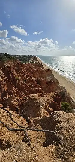 On the left, eroded red sandstone cliffs, with pine trees above. And on the right a bright blue sea lapping a light sany beach.