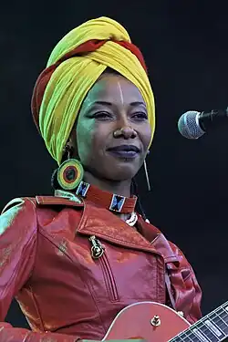 close-up of Fatoumata Diawara wearing a red leather jacket and colourful headwrap, standing onstage, holding an electric guitar, smiling