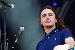 Against a black concert background punctuated by a hazy white light, a man in a blue shirt stands next to a pair of microphones.