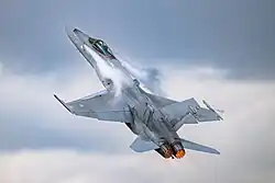 Jet fighter aircraft is seen against blue sky executing a pull-up, making it nearly vertical with contrail formed aft of the canopy