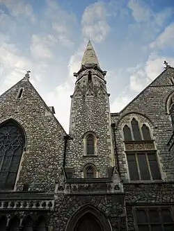 A picture of a church, built out of stone, with stained glass windows, viewed upwards from the street. Behind the church, a blue sky is lightly filled with white clouds.