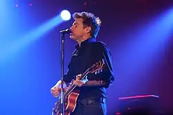 Bryan Adams wearing a black shirt and performing with his guitar under blue lights.
