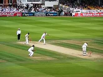 Men in cricket whites play upon a green grass cricket field amidst a stadium.
