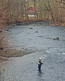 A wide muddy stream with some rapids surrounded by woods with bare trees and a red structure visible through them behind a bend at the rear. At the left are two men knee-deep in the water, holding fishing rods with a colored line attached