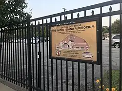 The plaque is hanging on a fence around the current parking lot. It contains the text in the description, along with a diagram of the façade of the former auditorium.