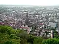 A view of Freiburg's old city taken from a hill. There are lush green trees in the foreground. In the middle ground, the Freiburg Minister is visible, as are older buildings. There are buildings out to the horizon, but the entire scene is very calm.