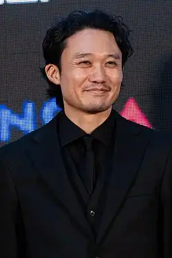 Fukunaga in three-quarter profile at press event wearing black suit and tie