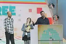 Three people, two women and a man, stand on an awards stage with a podium.