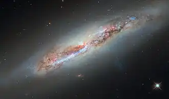 A spiral galaxy seen nearly edge-on. Its disk is filled with red and blue lights from star-forming nebulae and clusters of hot stars, respectively, as well as thick dark clouds of dust that block the strong white light from its centre. A faint, glowing halo of gas surrounds the disc, fading into the black background. A bluish plume of gas also extends from the galaxy’s core to the lower-right of the image.
