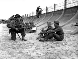 Juno Beach on D-Day, 1944. The barbed wire fence is crude and not very high. However, when combined with the steep, curving sea wall it slows down any attacker, giving time for the machinegun bunker (visible on the far left) to enfilade any attackers. Note the soldier in the background, forced to use a ladder.
