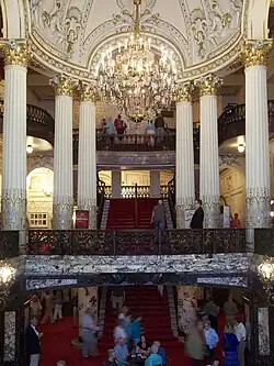 Interior of a grand theater lobby featuring red carpets, marble accents, towering white columns supporting upper balconies, and an elaborate crystal chandelier hanging from a decorated domed ceiling.