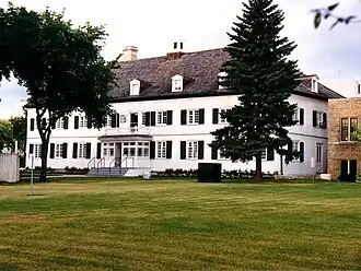White wooden, two-and-a-half-storey historic building with a hipped roof and dormers, set on a treed lot
