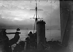 View from the cruiser HMS Sheffield as she sails on convoy duty through the waters of the Arctic Ocean. In the background are merchant ships of the convoy. The image was taken during the twilight of the arctic winter—the short time each day that the sun is seen during winter near the pole. In the foreground is the silhouette of a lookout using a telescope.