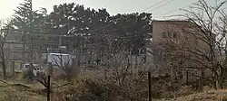 Panoramic view of the power station in autumn. On the left is an outdoor switchyard with numerous insulators, transformers, and metal structures. On the right is the pale pink, two-story HPP building. In the foreground are dry grass and bare tree branches. In the background is a wall of tall, dark green coniferous trees.