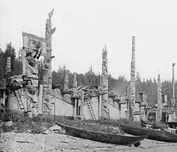 Haida longhouses with carved totem poles along the shoreline at Skidegate Inlet, British Columbia, with wooden canoes in the foreground, 1878.