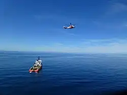 Red helicopter flying above a supply vessel during offshore support operations on calm sea.