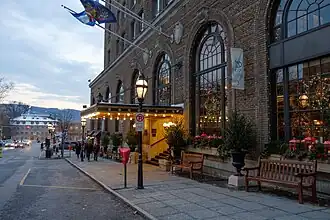 Street view of the hotel entrance, with warm lights under the canopy, holiday decorations, benches, and pedestrians walking along the sidewalk in the early evening.