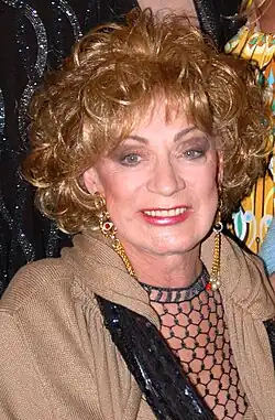 Holly Woodlawn smiles warmly while wearing gold earrings, a tan wrap, and a black sequined top with a netted neckline.