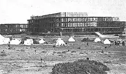 Black and white photo of a large partially constructed building with tents erected on the empty field nearby