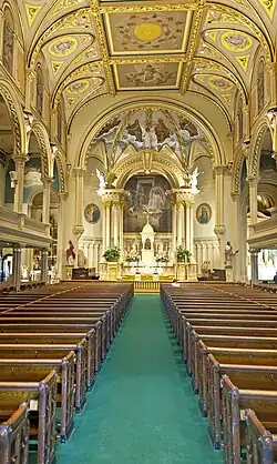 A green-carpeted aisle between two sets of wooden pews with a golden-colored decorated ceiling, arches over the side galleries and an arched, painted nave at the rear