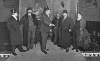 Photo of six white men standing, in Victorian business dress, four wearing hats; Edwardes, center, is speaking to Warde and gesturing.