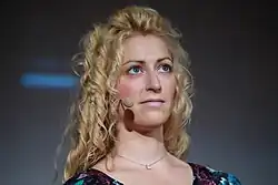 A woman with long curly blonde hair and blue eyes wears a headset microphone and a floral-patterned top while looking slightly upward