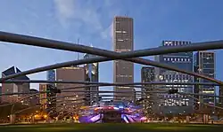 Evening view of a green lawn under a lattice with a lit metal bandshell against a backdrop of skyscrapers