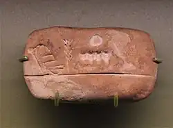 Uruk III administrative tablet from Jemdet Nasr. Ration account, here the reverse side which consolidates the data appearing on the front: the sign on the left combining the head and the bowl means "payment/expenditure" GU7, the stalk to its right means "barley" ŠE; on the right the numbers in the cereal counting system: the large circle worth 60 (N45) and the notches each worth 1 (N01). Ashmolean Museum.