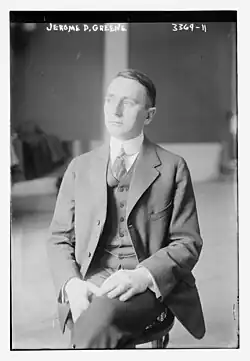 A black and white photo of Jerome Greene sitting on a wicker chair with his legs crossed at the knees, looking to the left, wearing a three-piece suit. Jerome D. Greene and 3369-11 are written at the top of the photo.