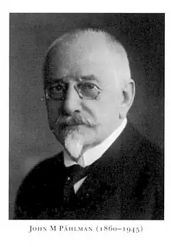 A black-and-white studio portrait of a bearded man with spectacles.