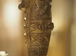 Close-up photograph of an ancient gilded bronze statuette, featuring detailed carved patterns and textures. Among the details are carved ram heads, an eagle with spread wings, and four-pointed stars.
