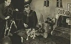 A sepia-coloured image of a film set - a small puppet (Mr. Prokouk) is being filmed sitting on the back of a miniature horse and cart. The director of the film, Karel Zeman, is standing next to the figures.