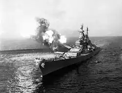 A black-and-white photograph depicting a large gunship sailing toward and slightly to the left of the camera. Guns of various size are visible on the ship, with smoke and flames visible from the turret No. 2 as the gun fires at an unseen target. The pressure from the gun fire has created a disturbance on the water surface
