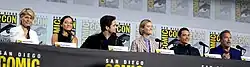 Linda Hamilton, Natalia Reyes, Diego Boneta, Mackenzie Davis, Gabriel Luna and Arnold Schwarzenegger seated at a table on a dais.