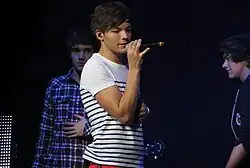 A young male wearing red trousers and a white t-shirt with black horizontal stripes sings into a microphone on a stage.