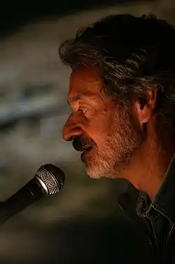A man with gray hair and a beard speaks into a microphone under warm lighting