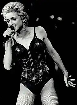 A blonde woman onstage with curly hair holding a microphone, wearing a black dress