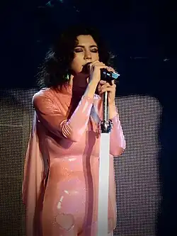 A young brunette woman singing into a microphone while playing keyboards. She is wearing a pink unitard and has a headpiece with a pair of purple Minnie Mouse-like ears.
