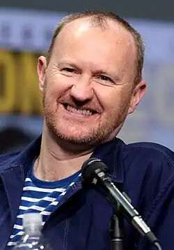 An image of a lightly bearded man, sitting at an interview panel with a microphone close to his face.