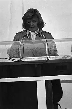 A woman with dark, shoulder-length hair wearing a light scarf and trench coat, speaking into microphones
