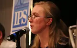 A woman with blonde hair and glasses speaks into a microphone