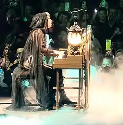 Lady Gaga sitting and playing the piano while wearing a veil-like black hooded cloak. A lantern is attached to the top of the piano.