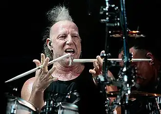 Mike Terrana sitting behind a drum kit onstage, spinning drumsticks between his fingers