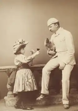Sepia photograph of a girl in Victorian clothing offering a wine glass to a man in a white suit, cap, and monocle, who is holding a live chicken.