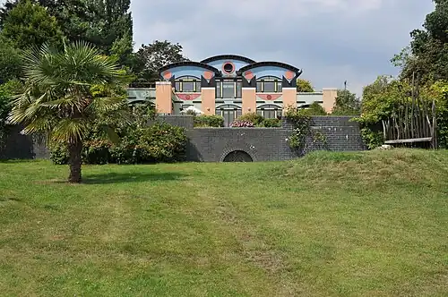 Postmodern house with playful Egyptian Revival influences, featuring stylized elements resembling Eye of Horus in upper facade, bold geometric shapes, and decorative red and black circles, surrounded by greenery and garden wall