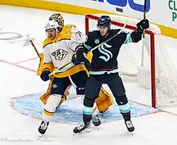 Ice hockey player in a Nashville Predators jersey holding his stick up next to an ice hockey player in a Seattle Kraken jersey