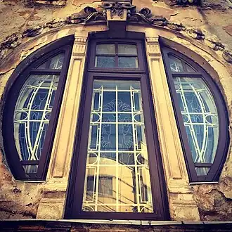 Tall arched window with three panes framed in dark wood, decorated with geometric white metal grilles, set in weathered stone wall