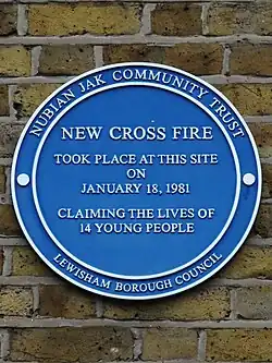 A blue circular plaque sits on the exterior of a brick building’s wall. The plaque reads "New Cross Fire took place on this site on 18 January 1981 claiming the lives of 14 young people." The outer ring of the plaque reads "Nubian Jak Community Trust; Lewisham Borough Council"