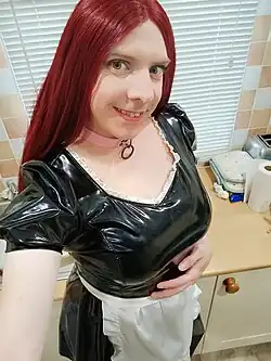 Selfie by a sissy smiling at the camera. He has long, red hair, and wears a black-and-white PVC maid uniform with breast forms underneath, a white apron, and a pink choker.