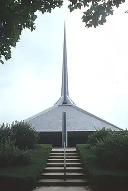 View of the church's roof and spire from the western entrance. Stairs ascend the berm to a driveway, behind which another stair climbs back down the berm. A ventilation shaft is visible in front of the church building, behind the stairs.
